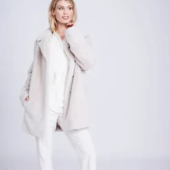 Woman wearing the Kennedy plush faux fur coat in pearl over white trousers, front view