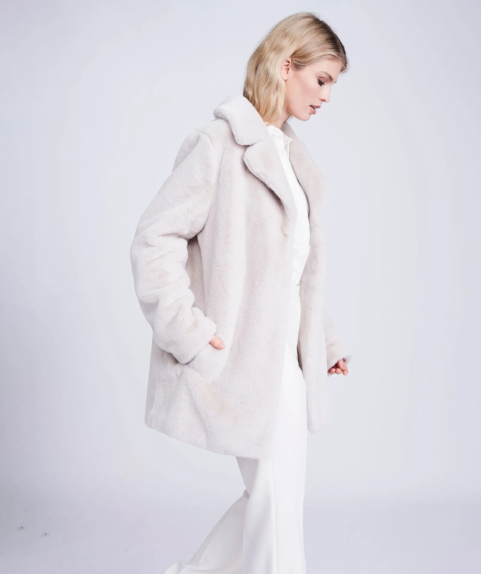 kennedy plush faux fur coat in pearl side Woman wearing the Kennedy plush faux fur coat in pearl, side profile, hands in pockets Back view of the Kennedy plush faux fur coat in pearl showing the wing collar Woman wearing the Kennedy plush faux fur coat in pearl over white trousers, front view